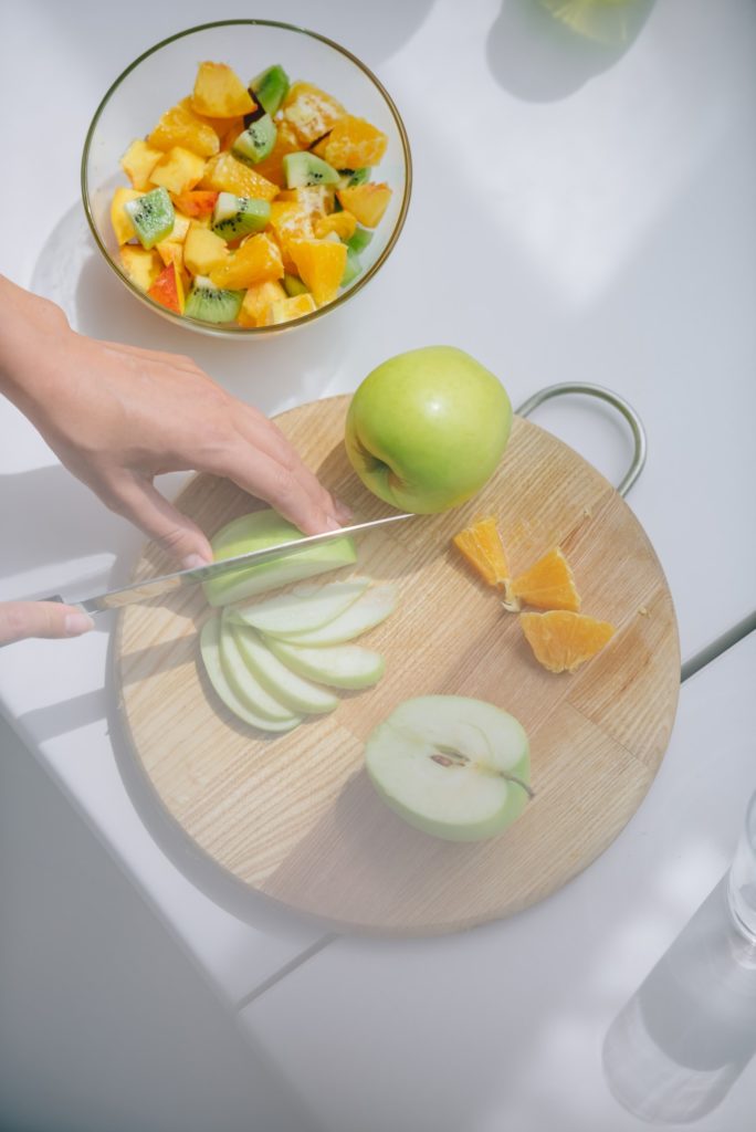 view of person's hands with knife cutting apple on cutting board to make fruit salad in bowl on side