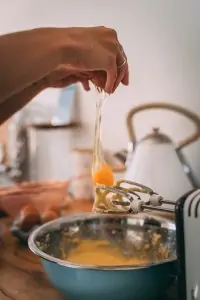 hand dropping egg yolk into silver mixing bowl with hand mixer on its side