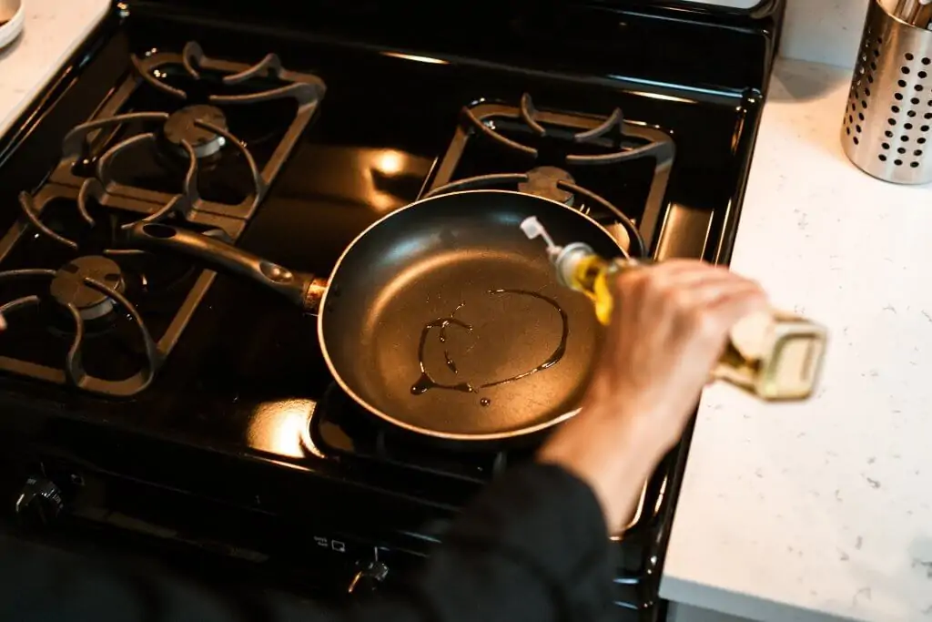 a person's hand drizzling oil from a glass jar into a black steel carbon pan on gas stovetop