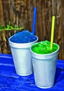 a bright blue and a bright green slushee with blue and yellow straws in white styrofoam cups on a table covered with a blue tablecloth in front of a wooden fence
