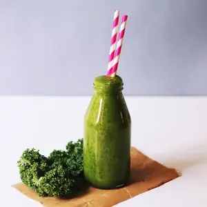 glass bottle with green juice in it with red and white straws and parsley on a white tablecloth
