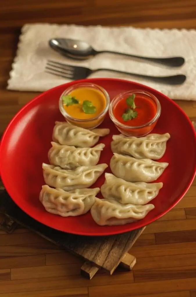 eight dumplings with two sauces on red plate on table next to white napkin with fork and spoon