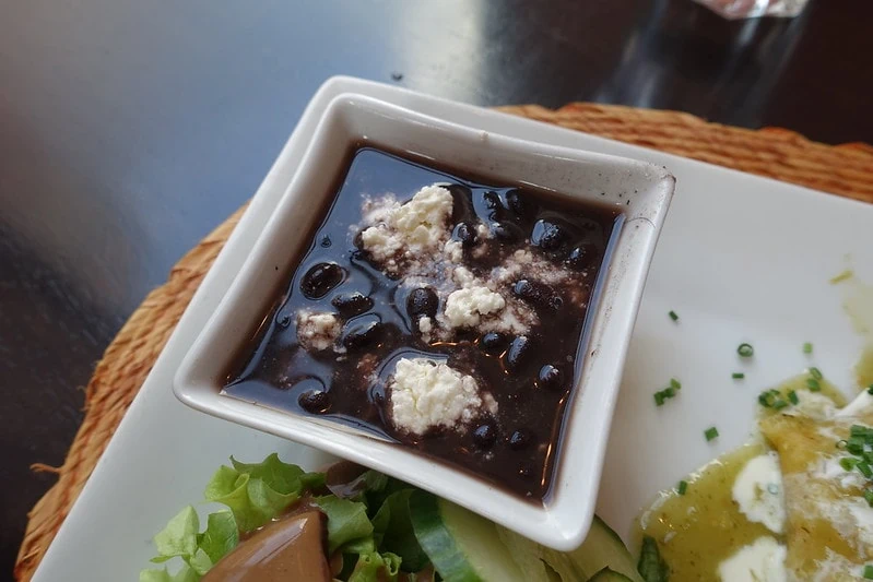 bowl of black beans in square white bowl on white plate with other food on side