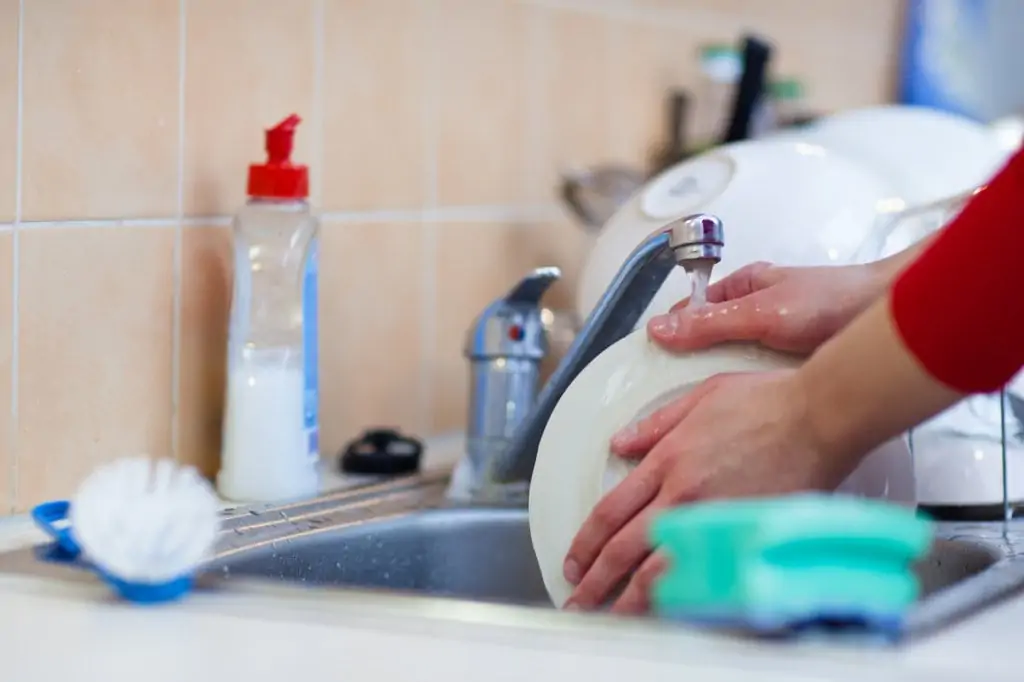 person's hands washing dishes in sink with brush and sponge on side of sink