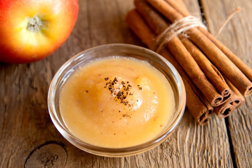 glass bowl of applesauce on wooden table with cinnamon sticks and an apple